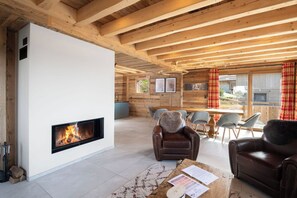 A modern interior with a fireplace, wooden beams, a dining area, and a view through large windows.
