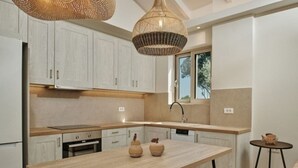 A modern kitchen with light wood cabinets, a central island, and a hanging light fixture. - Villa Danae Periyali (Zakynthos)