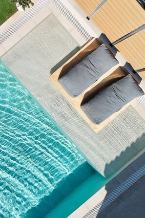 Una vista aerea di una piscina con acqua cristallina e blu, circondata da un solarium piastrellato e due sdraio con cuscini.