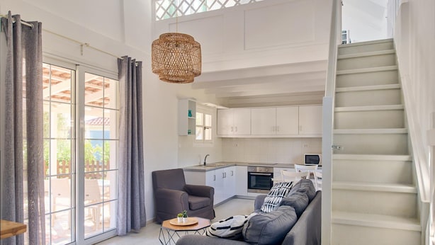A modern kitchen with white cabinets, a dining area with a grey sofa, and a staircase leading to an upper level. - Villa Cleo Periyali (Zakynthos)