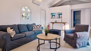 Ein modernes Wohnzimmer mit einem grauen Sofa, einem runden Couchtisch aus Holz und einem blauen Sessel. Ein Fenster bietet Blick auf Bäume, und an der Wand hängt ein Gemälde.