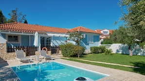 Ein Pool mit Liegestühlen und Sonnenschirm, umgeben von einem gepflegten Rasen und einem Haus mit einem Ziegeldach.