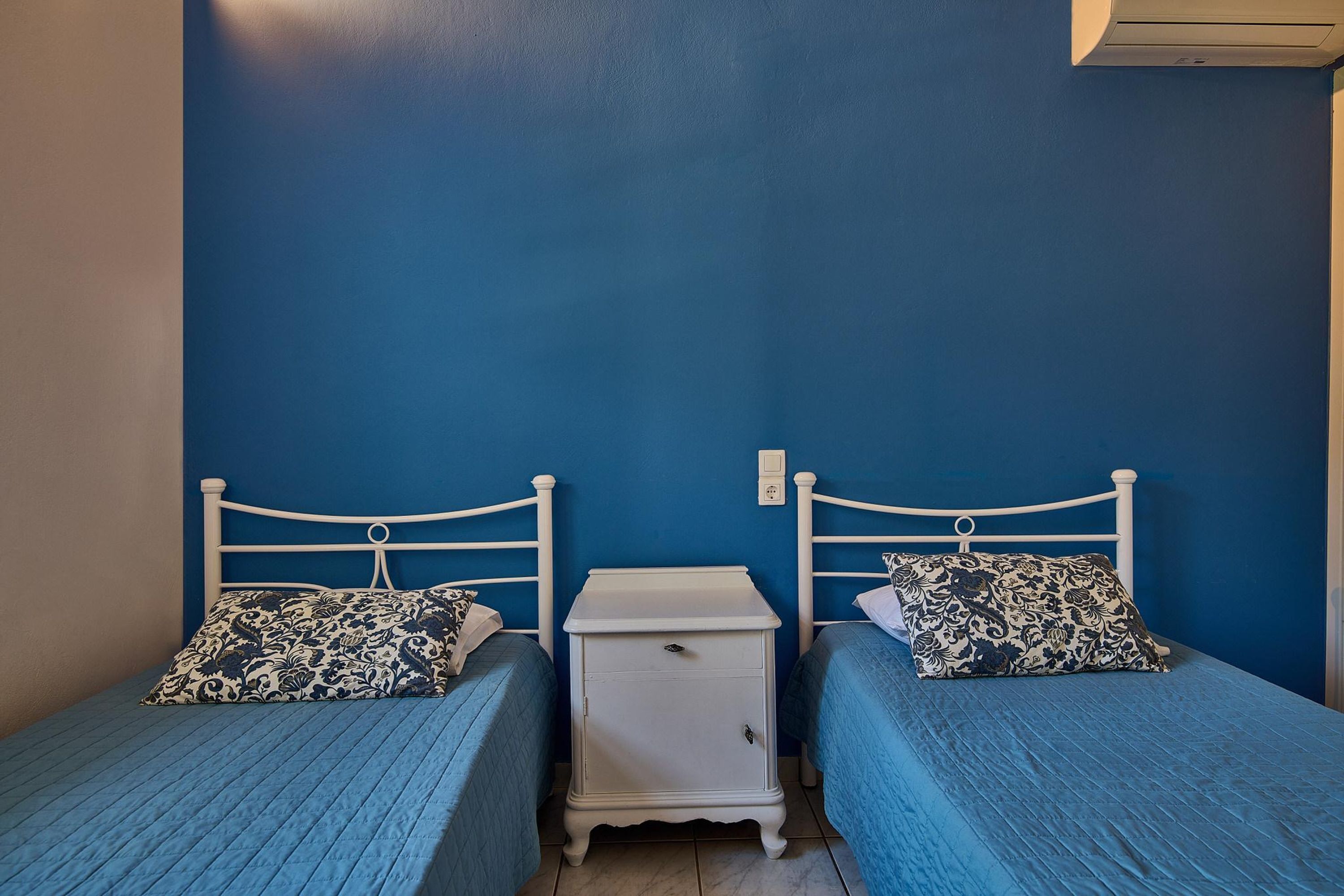 A bedroom with two single beds, each with a patterned pillow and a blue bedspread. There is a white bedside table between the beds.