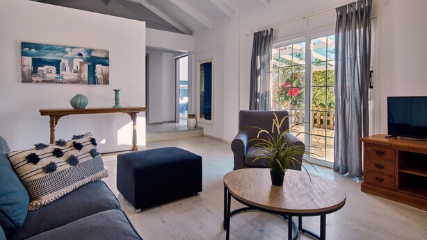 Ein modernes Wohnzimmer mit einer Couch, Hocker und einem Couchtisch mit Pflanze. An der Wand hängt ein Gemälde und ein Fernseher steht auf einem Holzkasten.