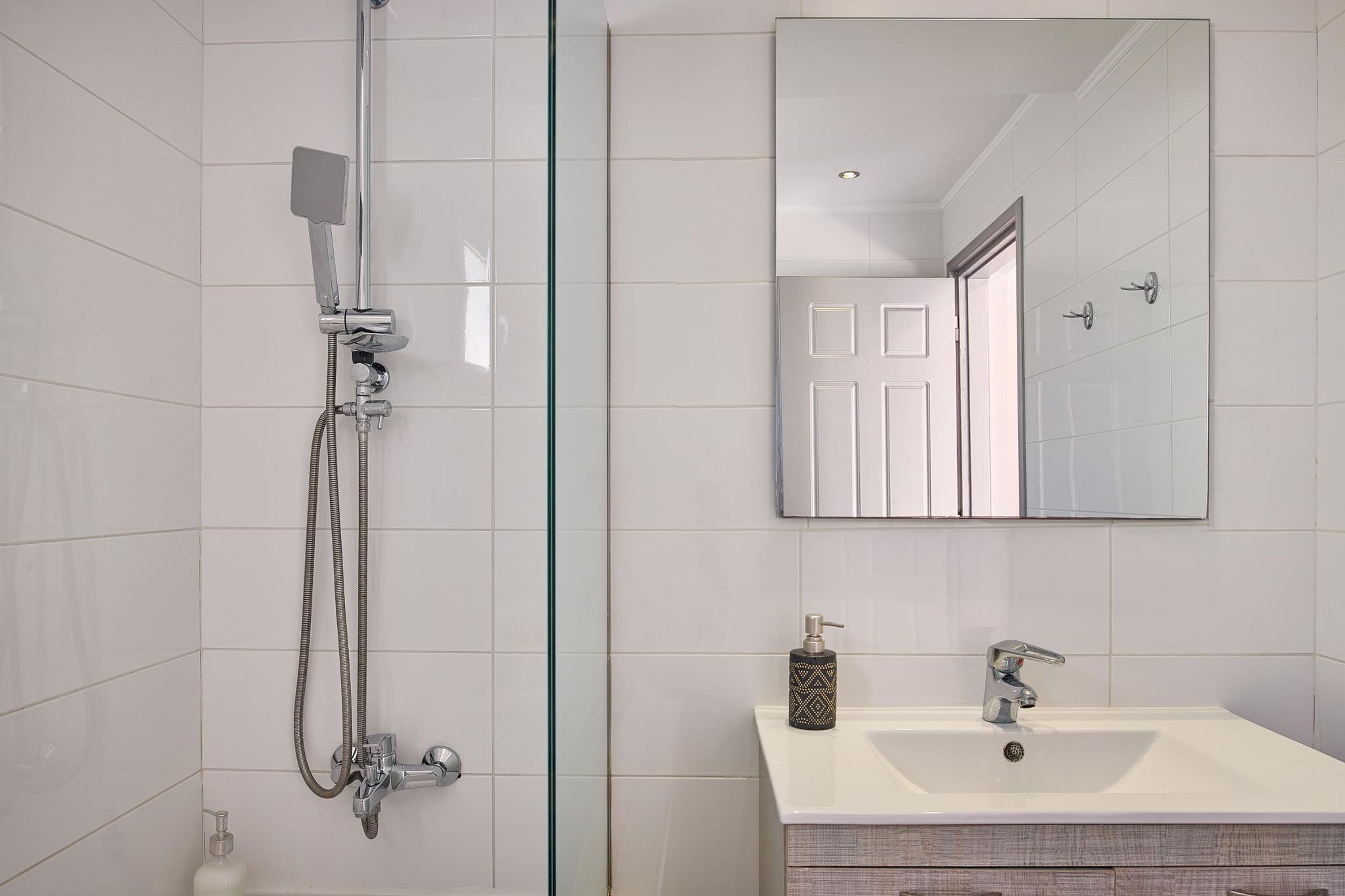 A bathroom with a glass shower enclosure, a white sink with a mirror above it, and a bottle of soap on the sink.