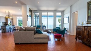 Un salón espacioso con un sofá grande, sillas color turquesa y un reposapiés rojo. Se ve una cocina al fondo, y grandes ventanales ofrecen vistas al mar.
