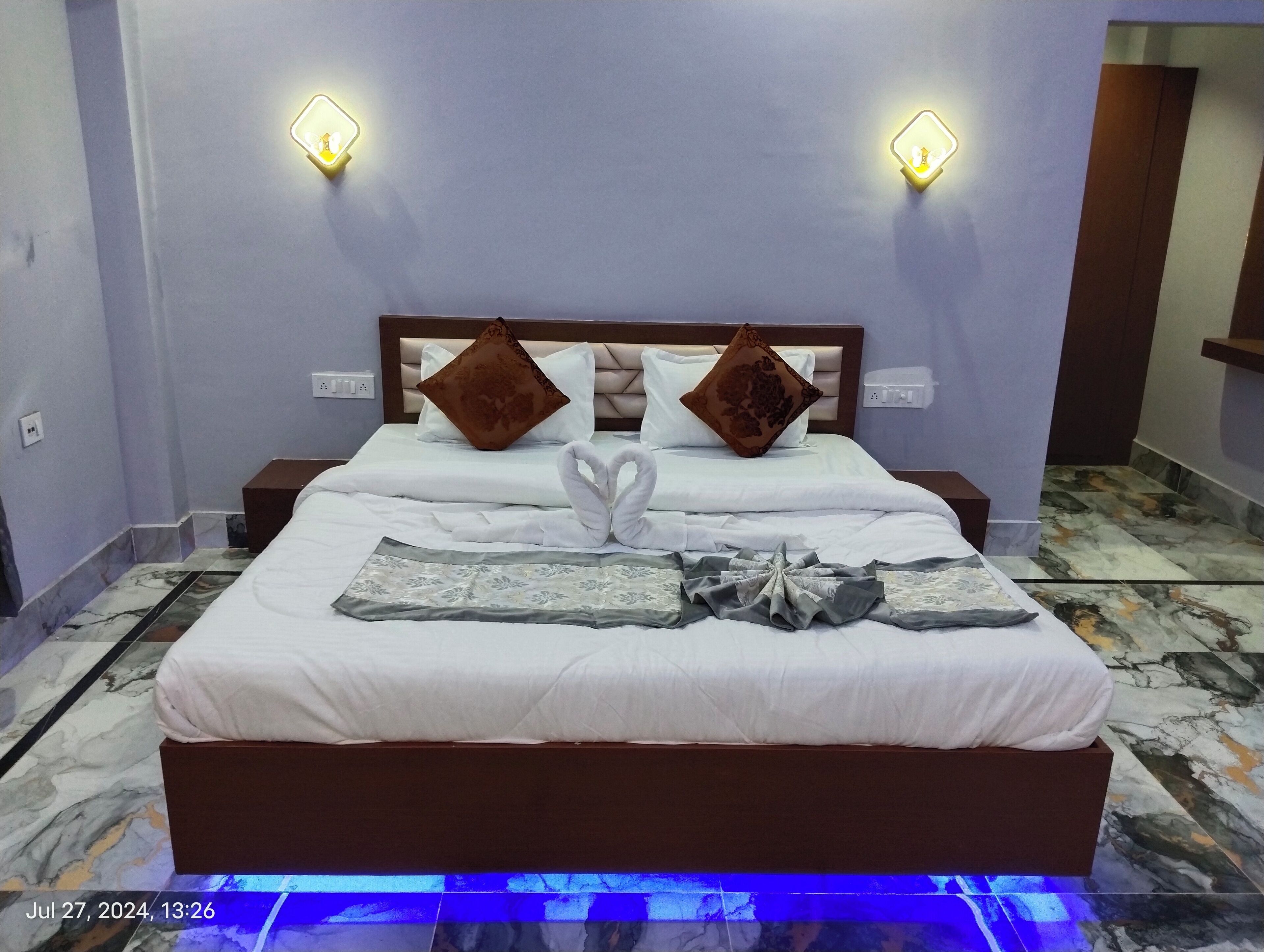 A neatly made bed with white linens, two decorative pillows, and a decorative item on the bed. There are two bedside tables with lamps, a marble-patterned floor, and a wall-mounted light fixture.