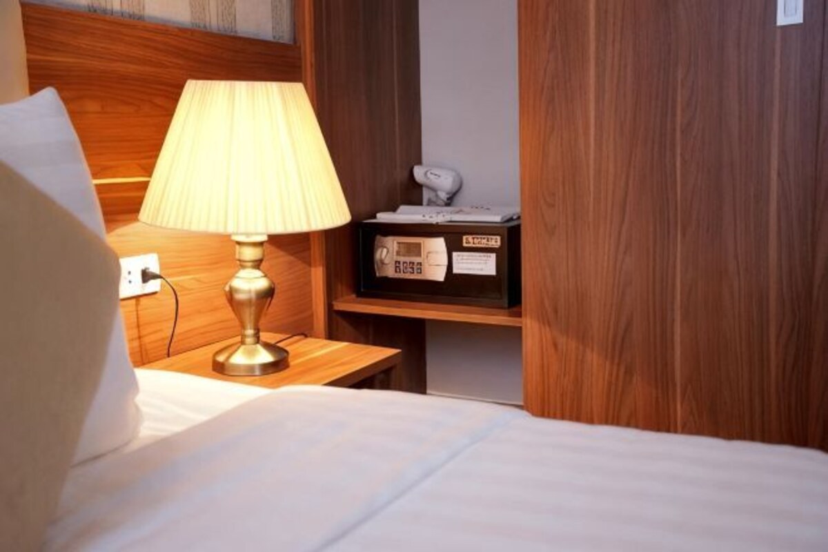 A hotel room with a bed, a bedside lamp, a wooden headboard, a nightstand with a lamp, and a shelf with a box and a clock.