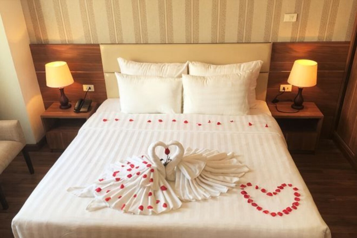 A hotel room with a neatly made bed, two bedside lamps, and a decorative swan and heart on the bedspread.