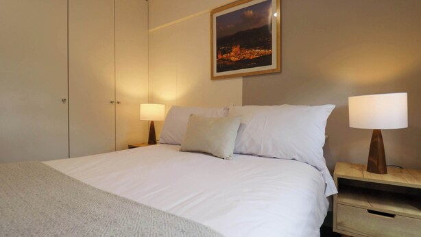 A neatly made bed with white linens and a patterned throw, a nightstand with a lamp, a framed picture on the wall, and a wardrobe. - w* | Bright 1BR in Historic Cusco w/ Balcony (Cusco)
