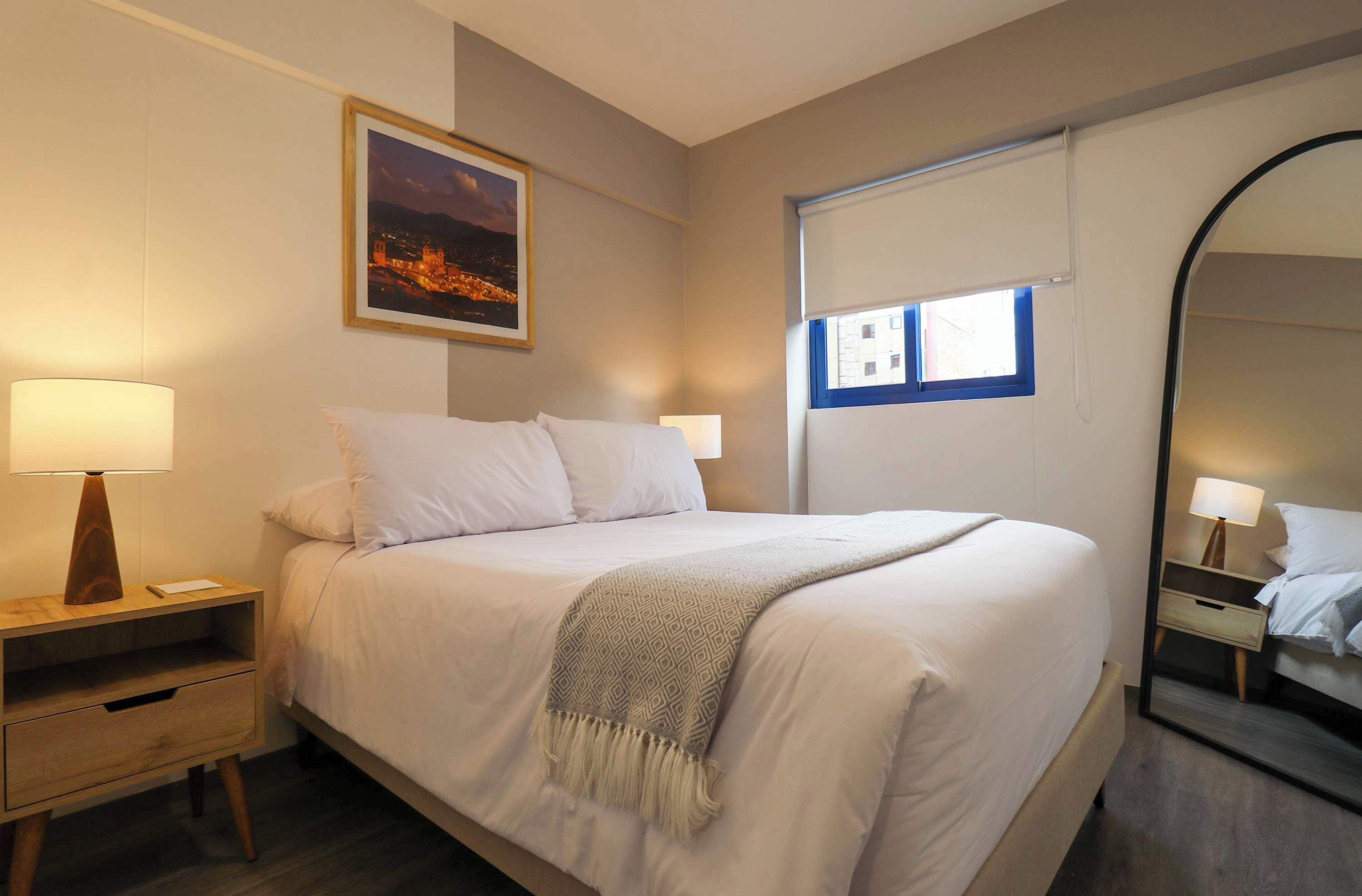 A neatly made bed with a patterned throw, bedside tables with lamps, a framed picture on the wall, and a window with blinds.
