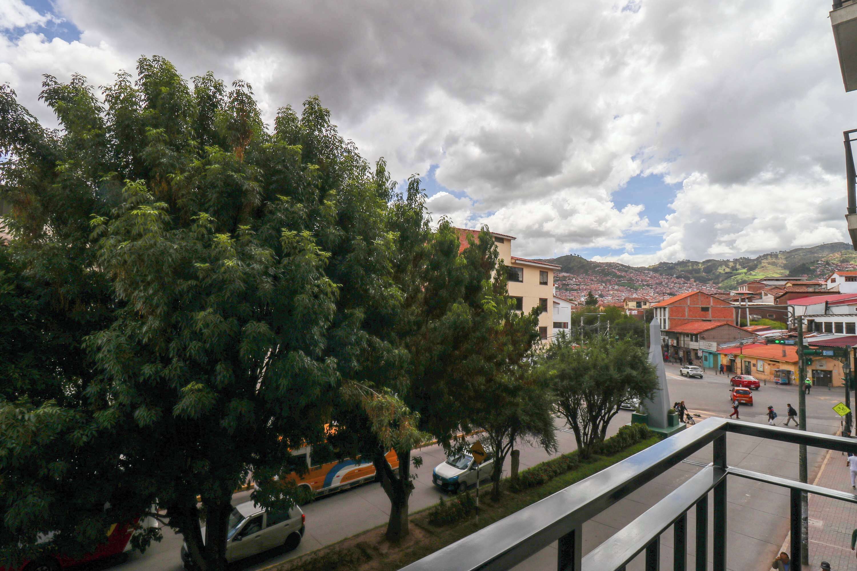 A view from a balcony overlooking a street with trees, buildings, and a distant mountain range.