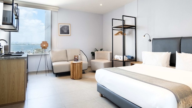 A modern hotel room with a large bed, a sofa, a small round table, a hanging clothes rack, and a view of the city. - w *|Grand 2BR w/ Lovely Terrace in Bocagrande (Cartagena de Indias)