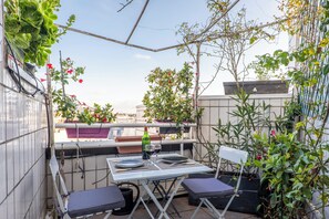 Ein kleiner Balkon mit Tisch für zwei Personen, umgeben von Blumentöpfen und Blick auf die Stadt.