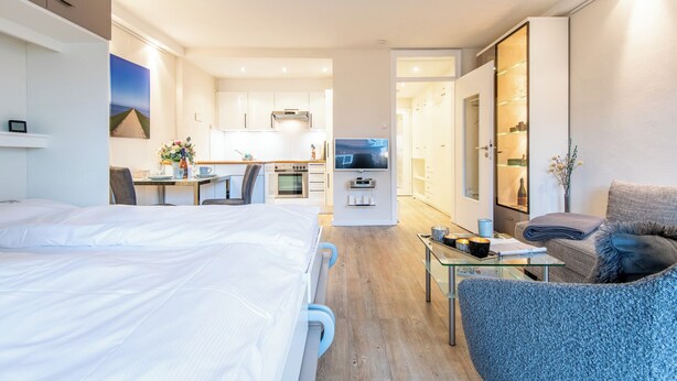 Ein modernes Hotelzimmer mit einem großen Bett, einer Sitzecke mit Glastisch, einer Küchenzeile und Blick auf die Stadt.
