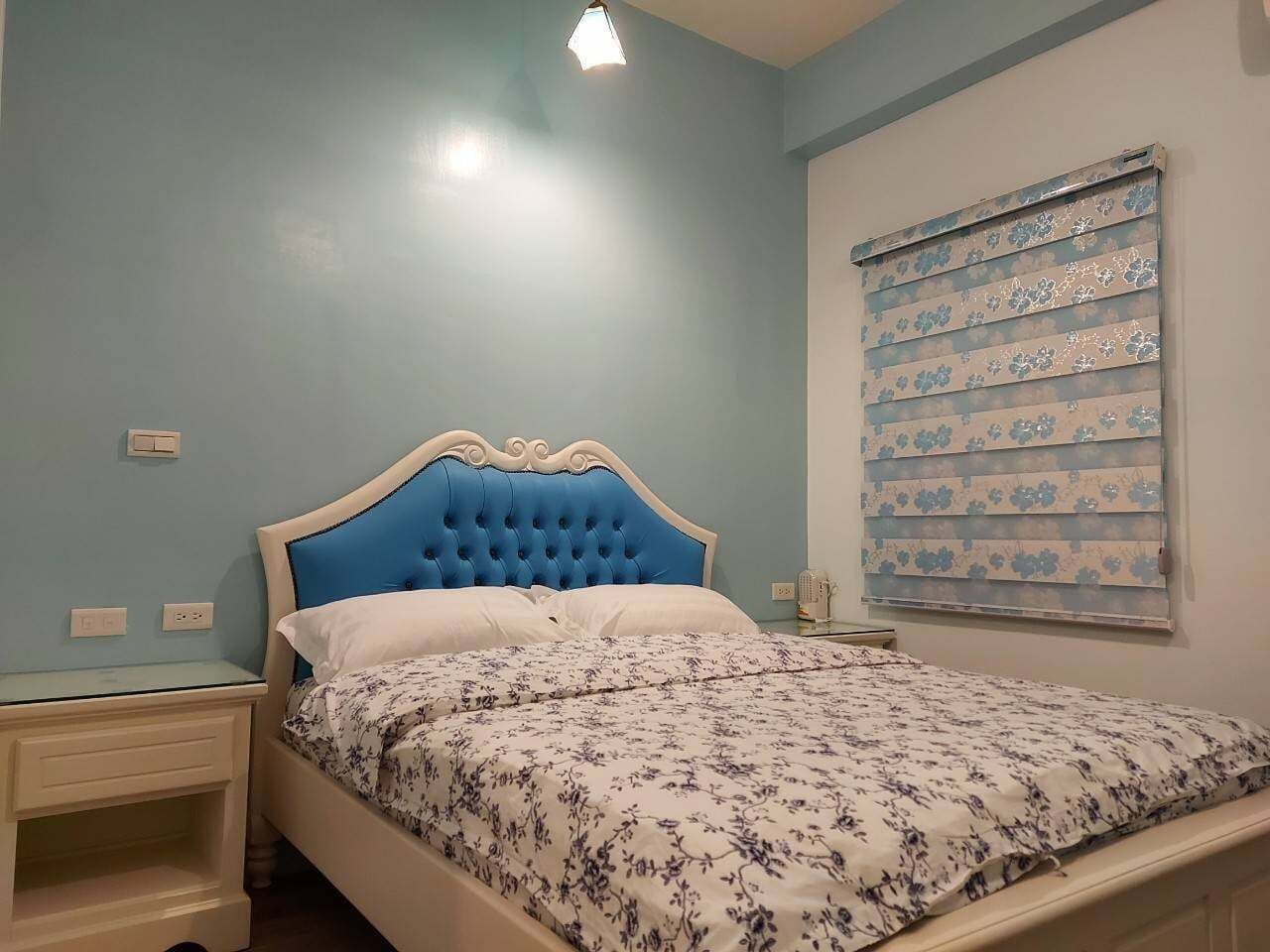 Une chambre à coucher comprenant un lit, des tables de chevet, une fenêtre avec des stores et un luminaire.