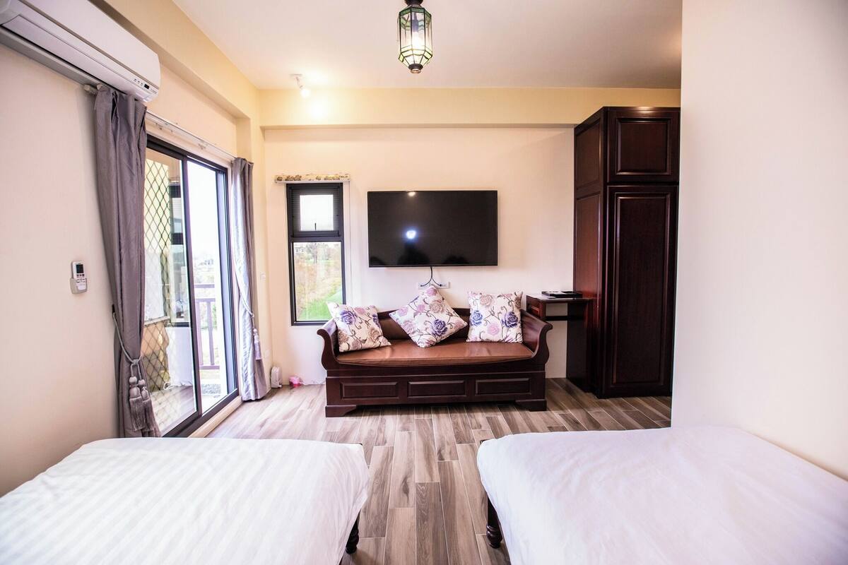 A hotel room with a sofa bed, two single beds, a TV mounted on the wall, and a wardrobe.