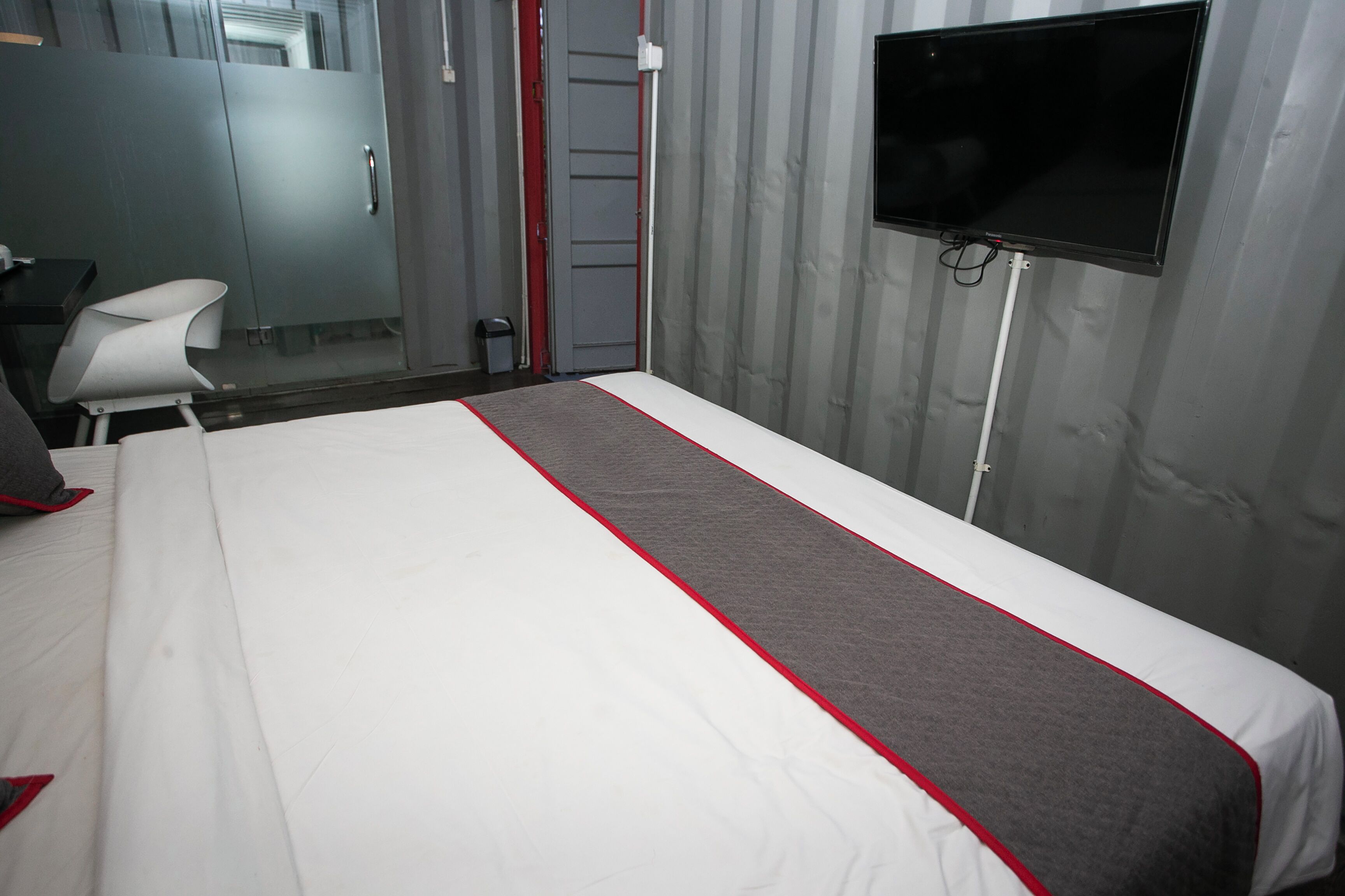 Quarto de hotel com uma cama, escrivaninha, cadeira, televisão e guarda-roupa.