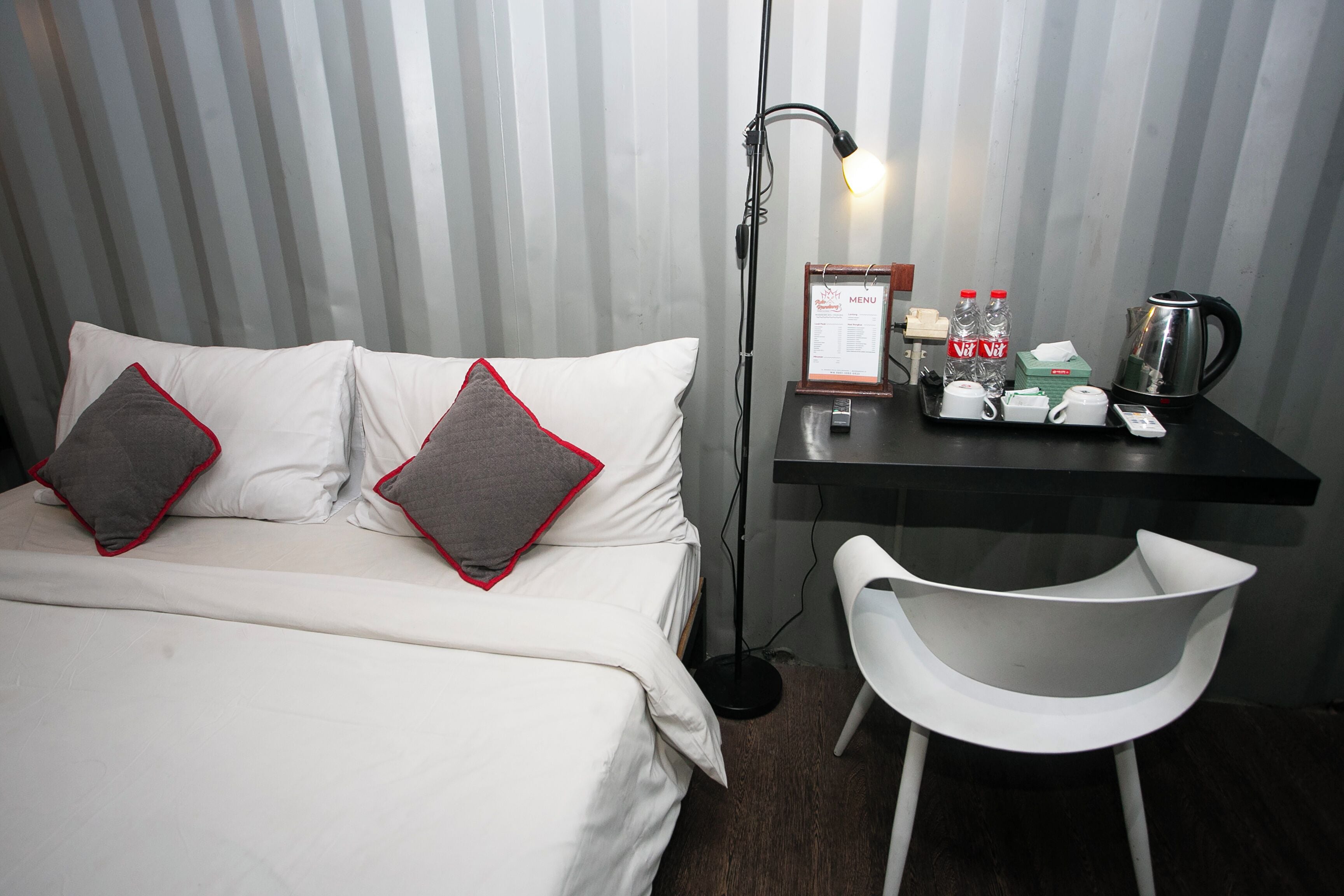 A hotel room with a bed, pillows, a desk with a lamp, a chair, a menu, and a kettle.