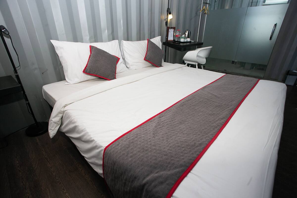 Una cama bien hecha con ropa de cama blanca y gris, un detalle decorativo rojo y dos almohadas con detalles en rojo y gris.