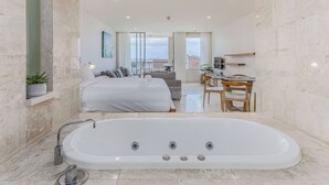 A modern bathroom with a large bathtub, a sofa, and a dining area visible through an open door.