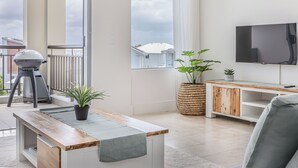 A modern living room with a flat-screen TV, a wooden coffee table, a grill, and a large window with curtains.