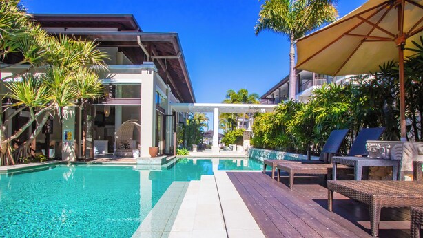 A clear swimming pool with a wooden deck and lounge chairs, surrounded by tropical plants and a modern building.