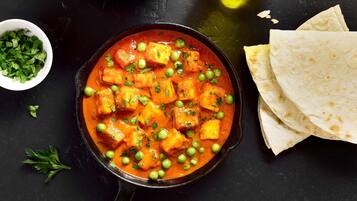 Un tazón de curry de paneer con guisantes y hierbas, acompañado de pan plano y un pequeño tazón de hierbas picadas.