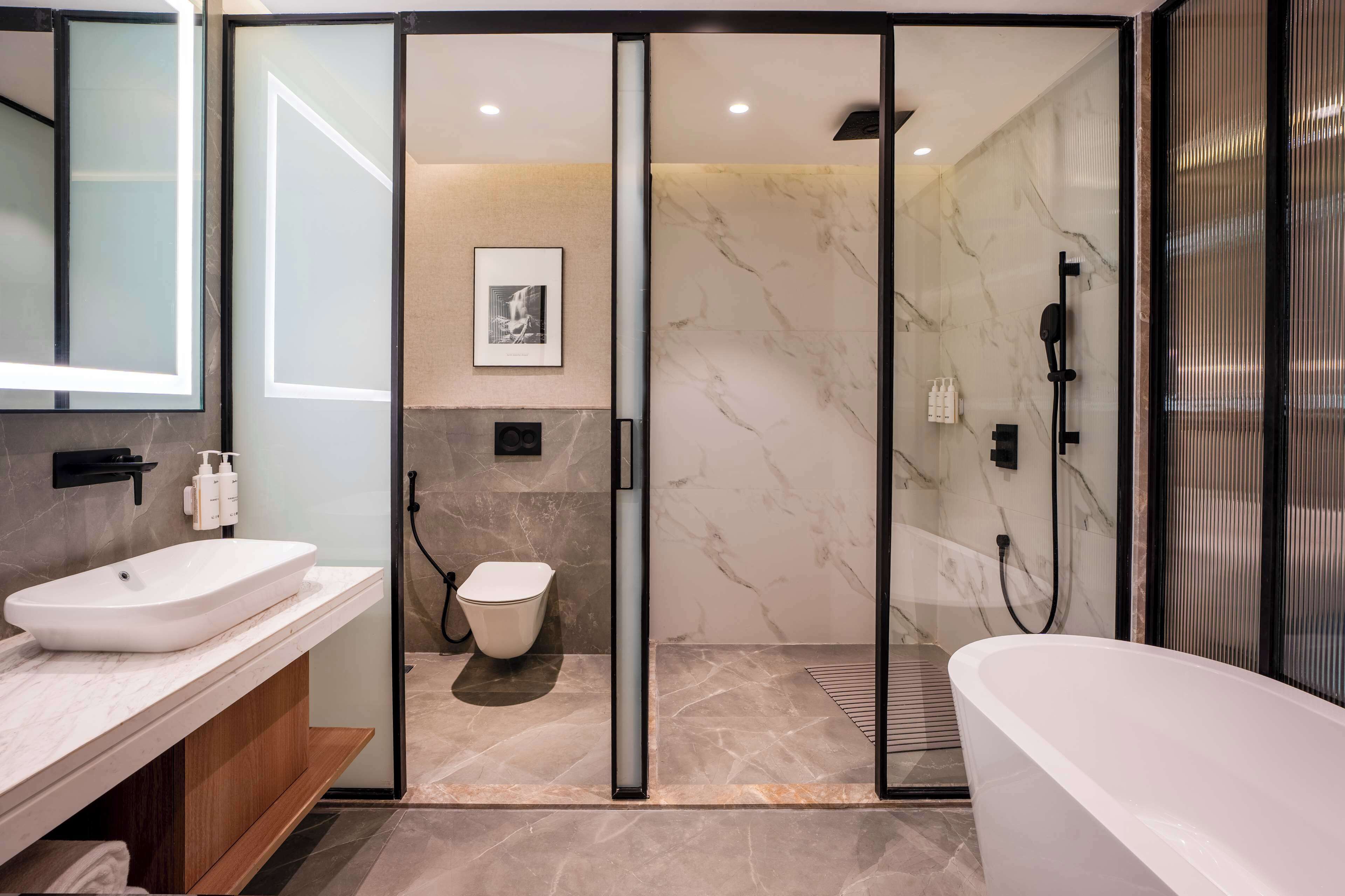 Een moderne badkamer met een wit wastafelmeubel, toilet en ligbad, marmeren wanden en een glazen douchecabine.