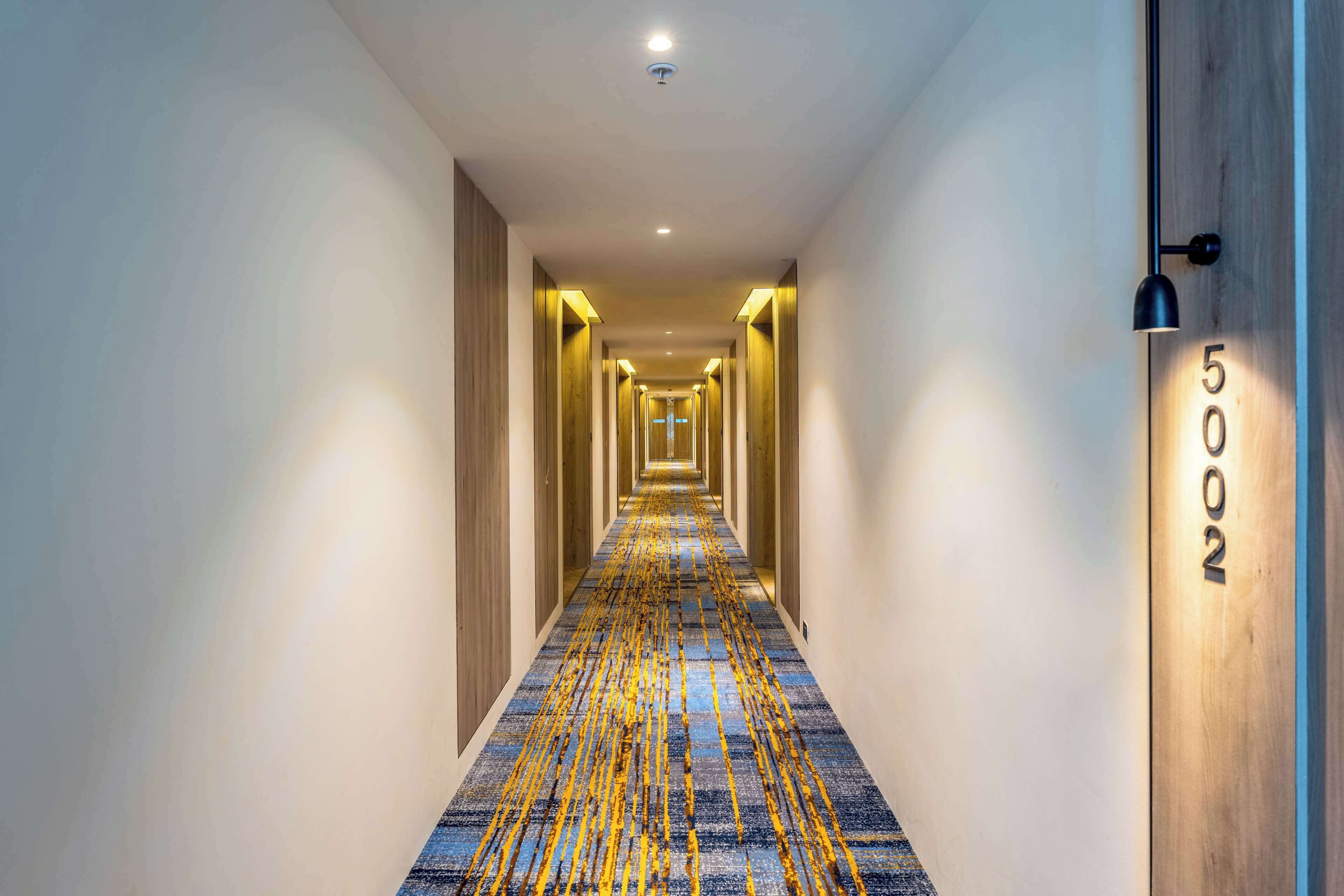 Um corredor de hotel com carpete estampado, portas de madeira e uma placa de número na parede.