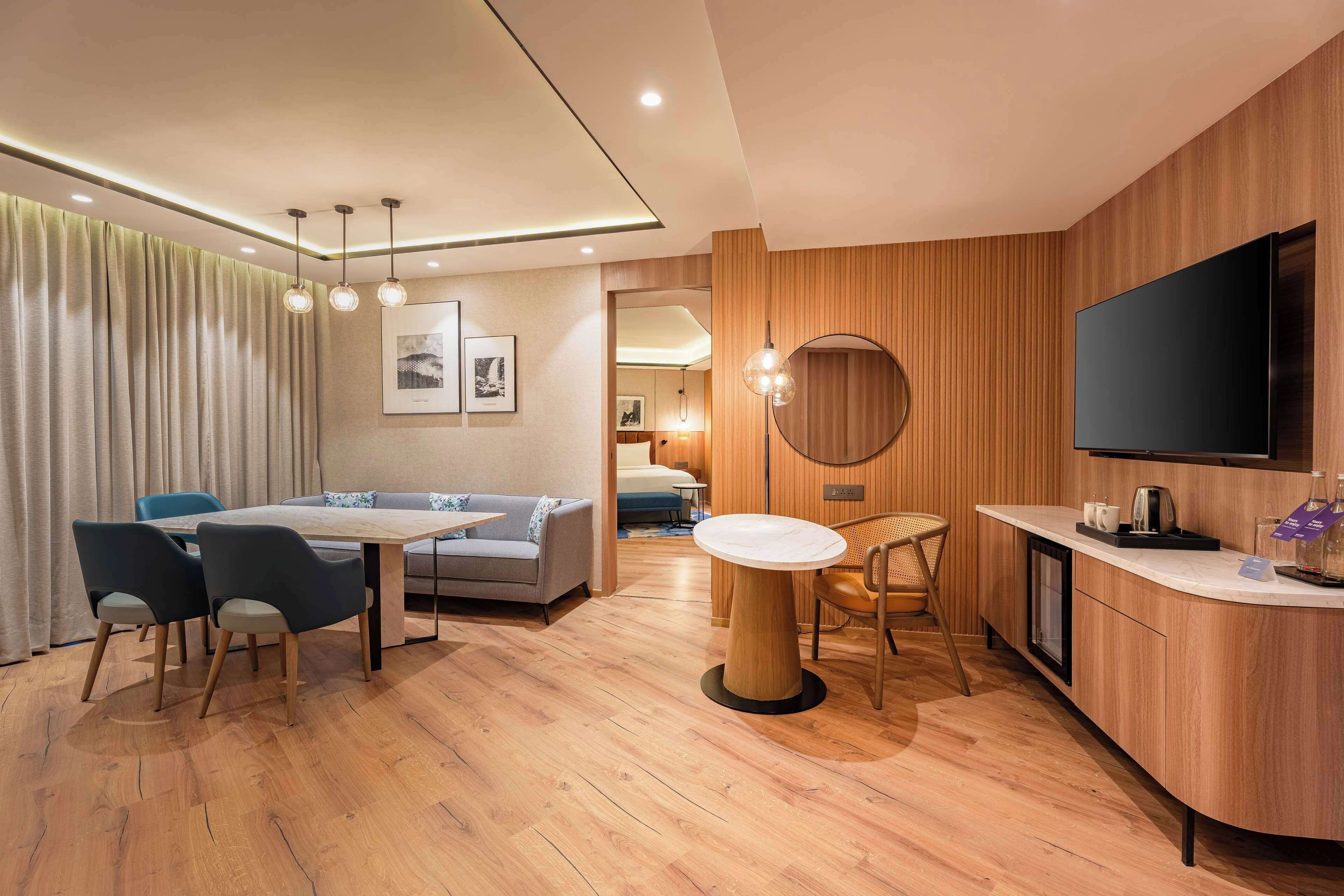 A modern hotel room with a wooden floor, a dining area with a table and chairs, a sofa, a round mirror, a flat-screen TV, and a kitchenette.