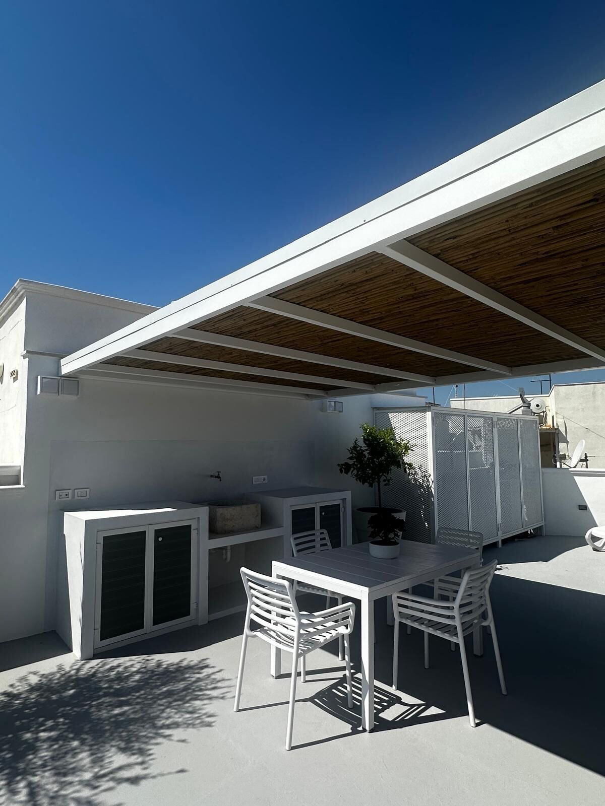 Eine Terrasse im Freien mit Tisch und Stühlen, einem Blumentopf und einem Gebäude mit weißem Anstrich und Holzdecke.