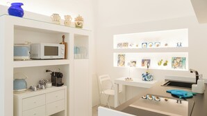A modern kitchen with white cabinetry, a microwave, a kettle, and a sink. There are shelves with decorative items and a chair. - FalcoMarino (Polignano a Mare)