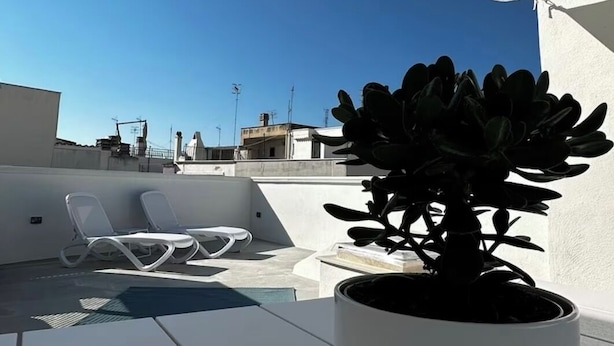 A rooftop terrace with a potted plant, white lounge chairs, and a clear blue sky.