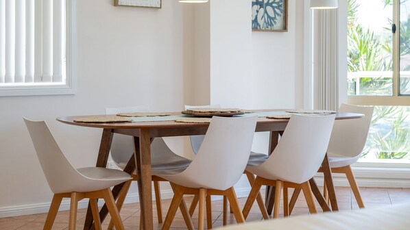 A dining area with a round wooden table, six white chairs, and a view of greenery outside. - Adriatic 4 in Tuncurry (Tuncurry)