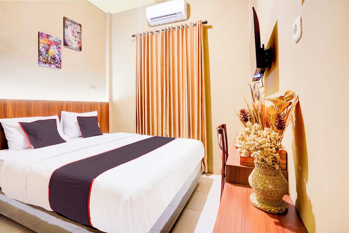 Quarto de hotel com uma cama grande, cabeceira de madeira, ar condicionado fixado na parede, cortina e um vaso decorativo com flores secas.
