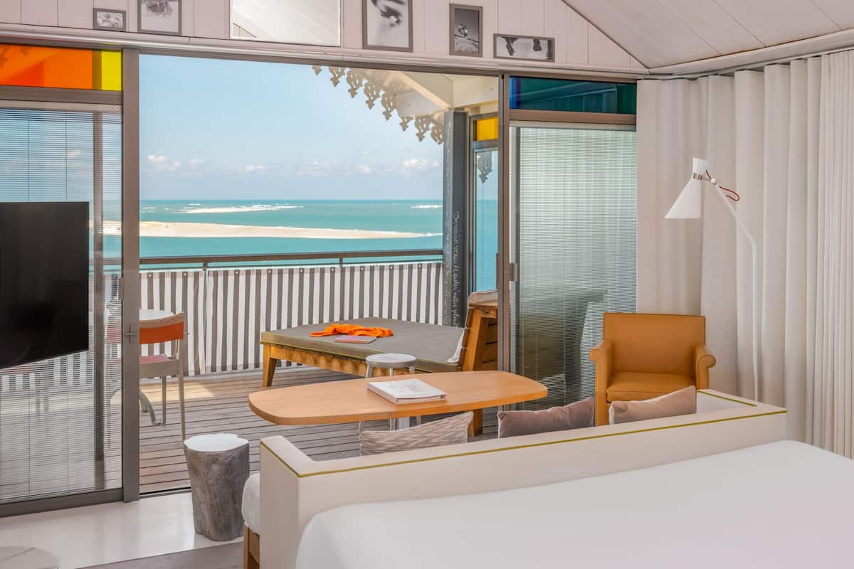 A room with a bed, a table, a chair, and a TV. There is a balcony with a view of the beach and ocean.