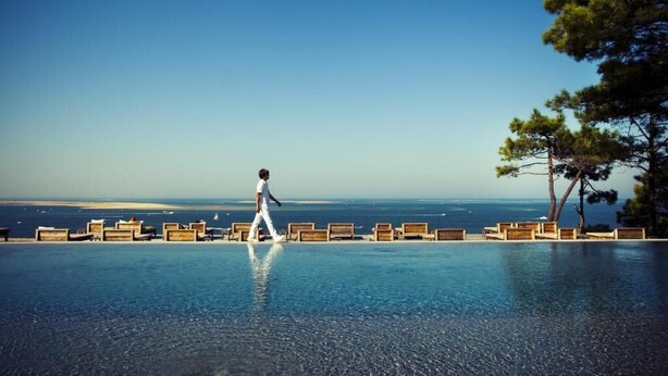 A person walking along a poolside with a view of the ocean and beach in the background. - La Co(o)rniche (La Teste-de-Buch)