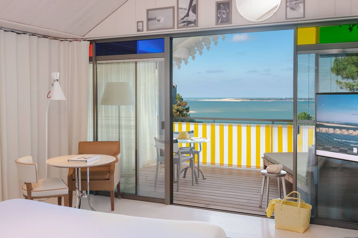 A hotel room with a large sliding glass door leading to a balcony with a view of the sea. There is a bed, a table with chairs, and a lamp.