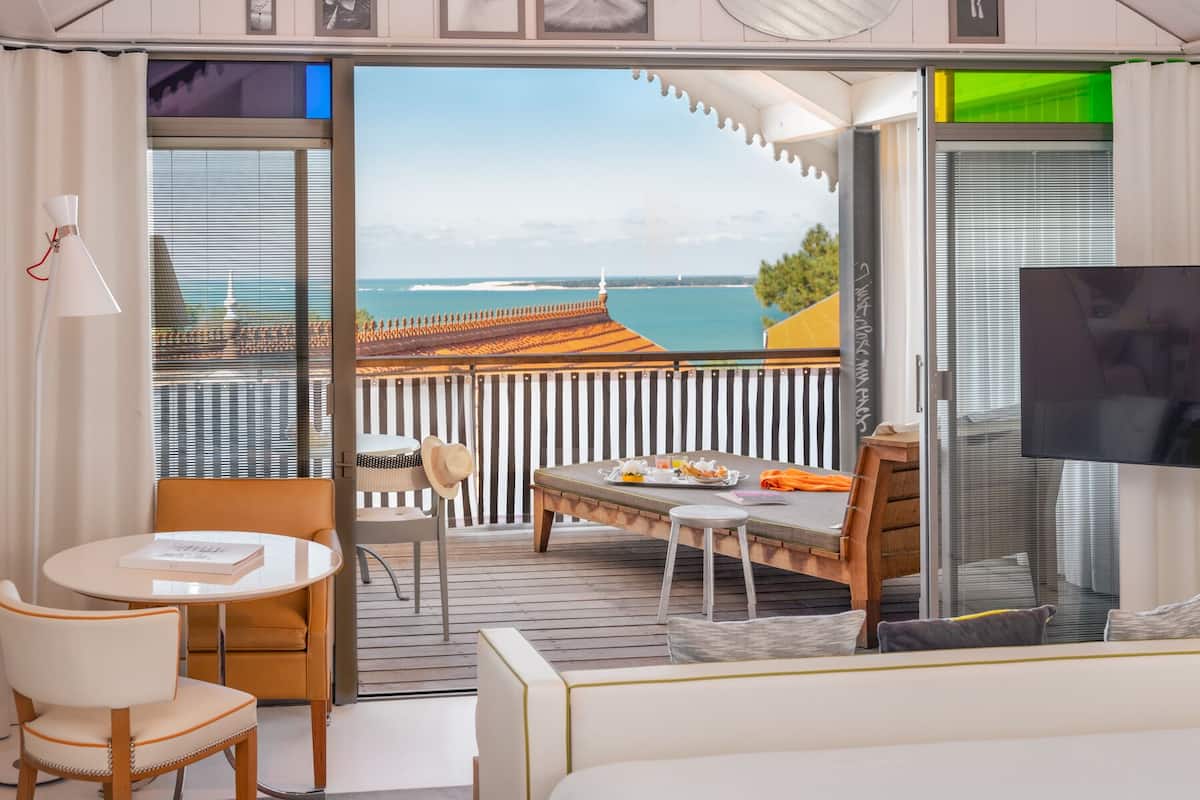 A hotel room with a balcony, a dining table, a sofa, and a TV.