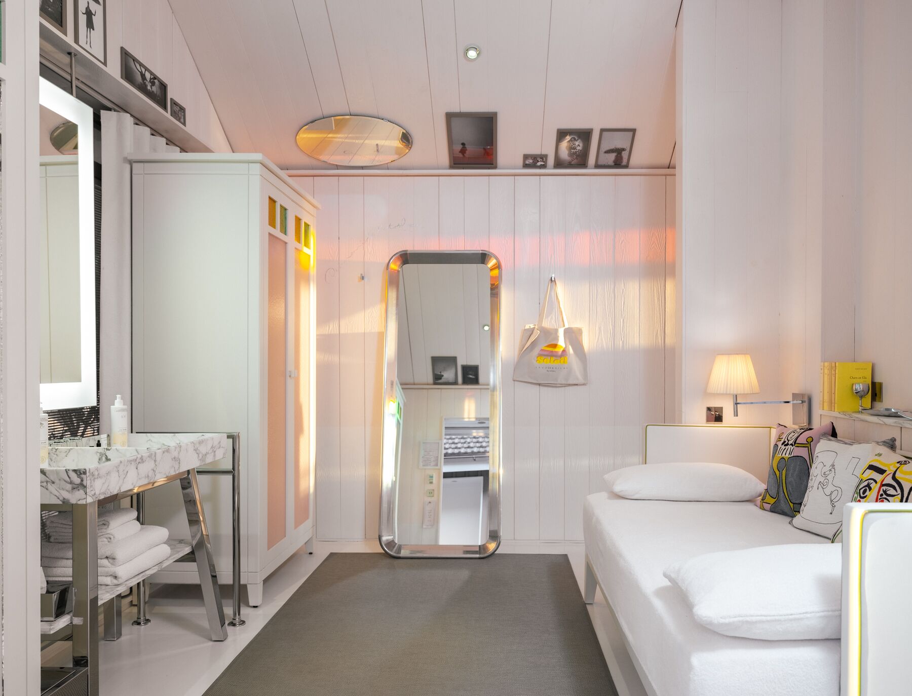 Una habitación de hotel moderna con tocador de mármol, un espejo grande, una cama con almohadas coloridas y una mesita de noche con lámpara.