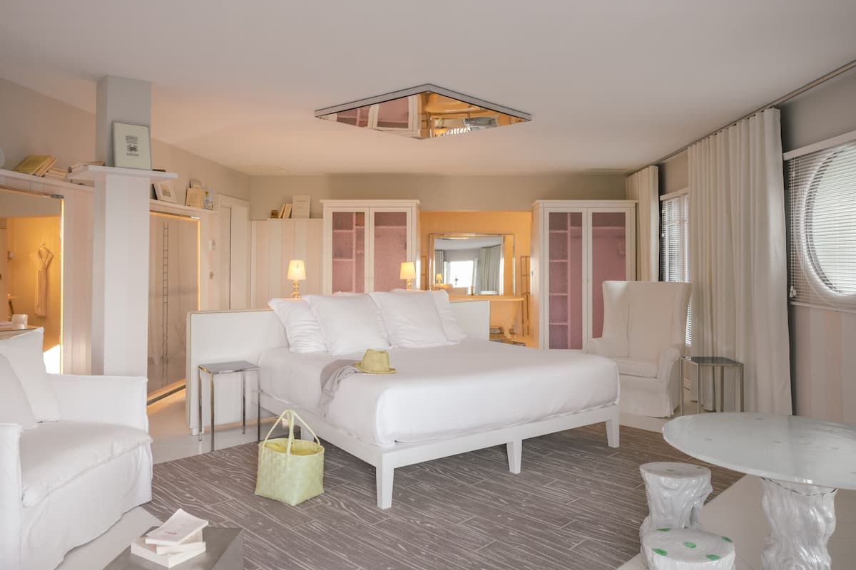 A spacious hotel room with a large bed, a sitting area with a chair and a table, a wardrobe, and a bathroom with a shower and a mirror.
