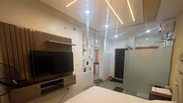 Habitación de hotel moderna con una cama grande, un televisor de pantalla plana, sistema de sonido integrado, cabina de ducha de vidrio y un pequeño rincón de descanso con un ventilador.