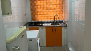 Una cocina compacta con armarios blancos y anaranjados, una estufa a gas, un lavamanos y una lavadora pequeña.