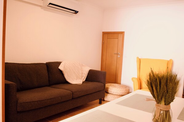 A living room with a brown sofa, a wooden door, a yellow armchair, and a vase of wheat.