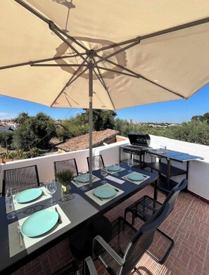 Een terras buiten met een eettafel, een barbecue en een grote parasol voor de schaduw.