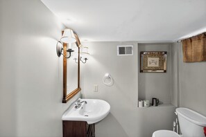 Ein Badezimmer mit einem weißen Waschbecken, einem Spiegel, einer Wandleuchte, einem gerahmten Bild und einer Toilette.