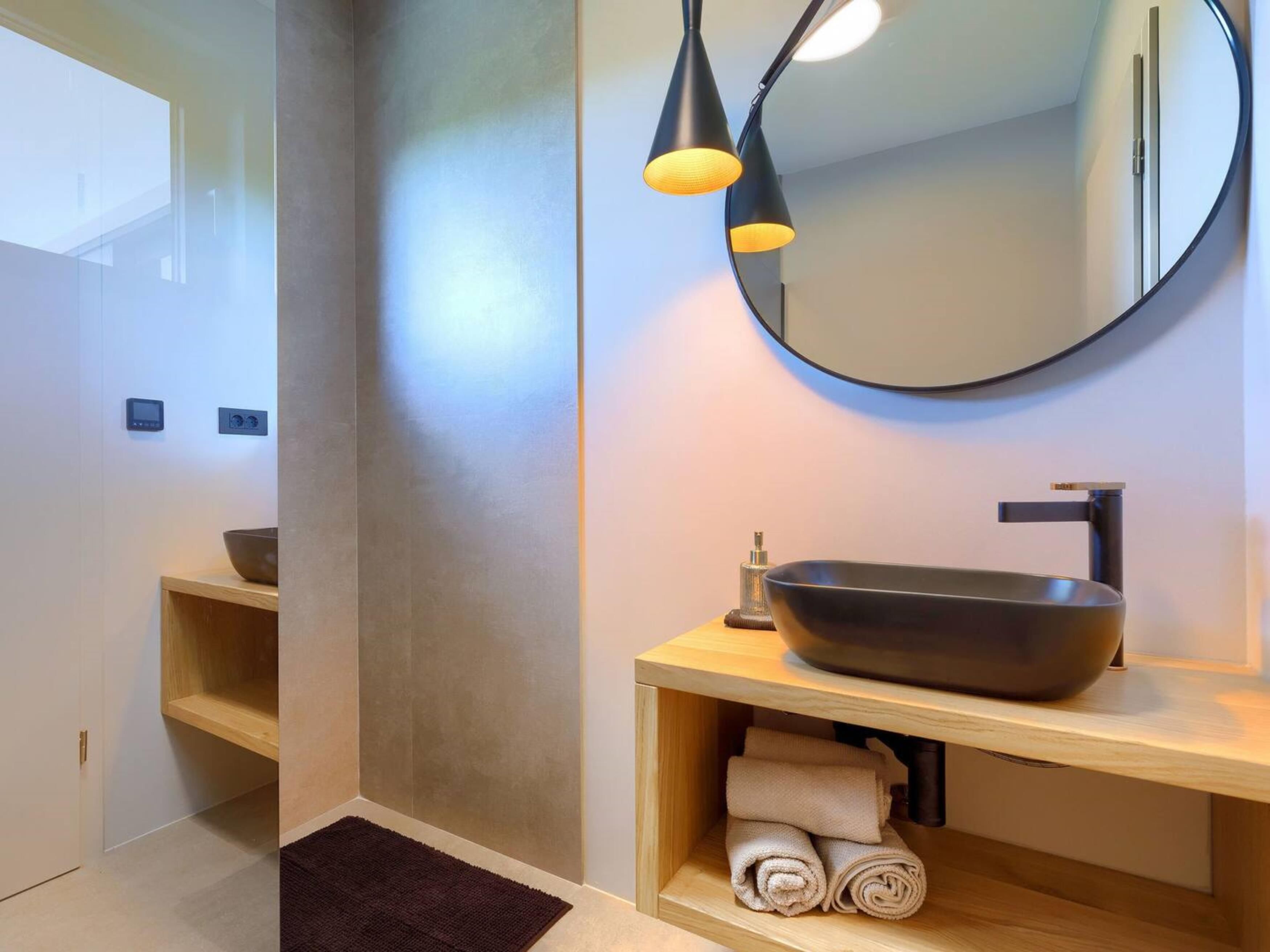 Une salle de bain moderne avec un grand miroir rond, un lavabo noir sur colonne et un meuble-lavabo en bois.