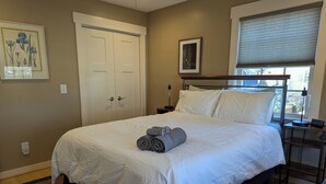 A neatly made bed with rolled towels, a bedside table with a lamp, a window with blinds, and a door with a coat rack. - Back Alley Apartment, walk Downtown and Duke. (Durham)
