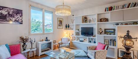 Ein modernes Wohnzimmer mit einem Glastisch, einem rosa Sofa, einem weißen Sessel und einem Bücherregal voller Bücher und Dekoration.